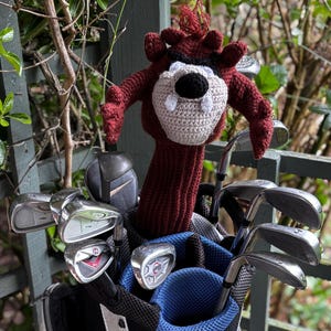May include: A golf club bag filled with golf clubs. A burgundy and beige crocheted golf club head cover shaped like a cartoon character is visible. The golf bag is black and blue.