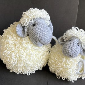 May include: Two crocheted sheep tea cozy sets. The larger sheep is white with a gray face and is covering a teapot. The smaller sheep is white with a gray face and is covering a teacup.