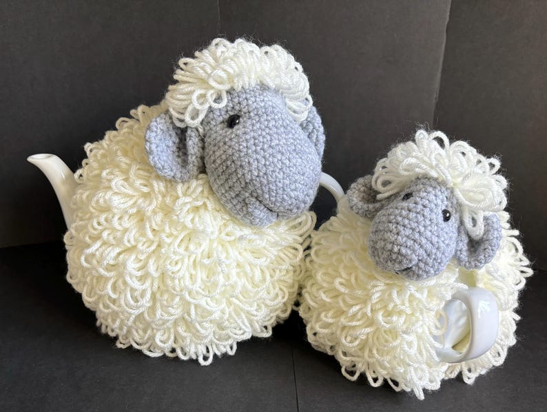May include: Two crocheted sheep tea cozy sets. The larger sheep is white with a gray face and is covering a teapot. The smaller sheep is white with a gray face and is covering a teacup.