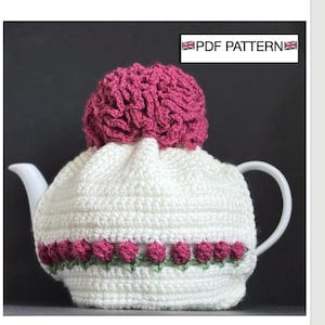 May include: A white crocheted tea cosy with a decorative row of pink flowers and green leaves. The cosy is topped with a textured, pink crocheted element. The text "PDF PATTERN" is displayed in a white box.
