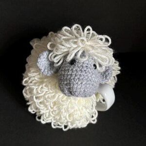 May include: A white crocheted sheep mug cozy with a gray crocheted sheep head and black eyes.