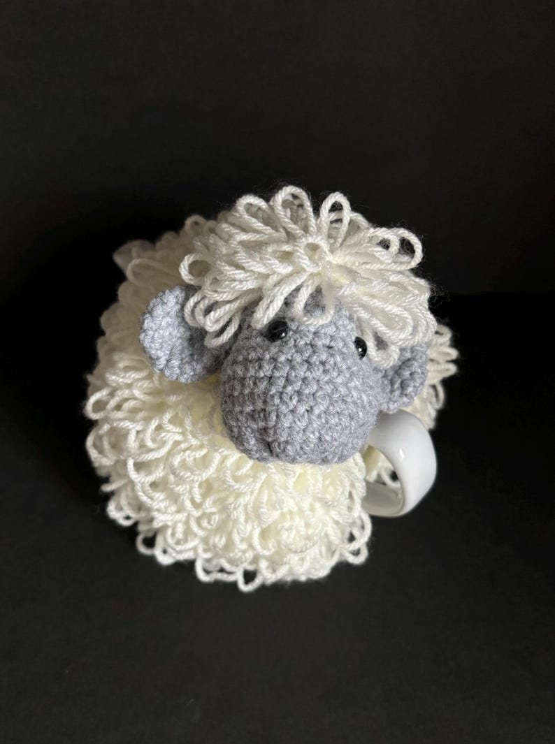 May include: A white crocheted sheep mug cozy with a gray crocheted sheep head and black eyes.
