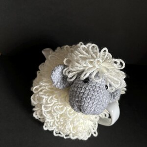 May include: A crocheted white sheep mug cozy with a gray sheep face. The sheep has a fluffy white wool head and a gray face with black eyes.