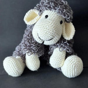 May include: A handmade crochet sheep doll with a smiling face. The sheep has a white head, legs, and ears, with gray curly wool and black eyes. The doll is sitting on a dark surface.