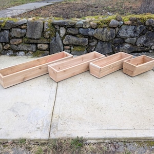 May include: Four unfinished rectangular wooden planters of varying sizes are arranged on a concrete surface. The planters are made of light-colored wood and are empty. A stone wall with moss and greenery is in the background.