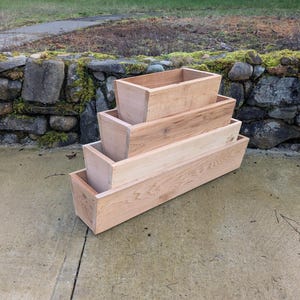May include: Four wooden planters of different sizes, arranged in a tiered formation. The rectangular planters have a natural wood finish, displaying the wood grain. The setup is on a concrete surface, with a stone wall and greenery visible in the background.