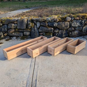 May include: Five unfinished rectangular wooden planters of varying sizes. The planters are made of light-coloured wood and are empty. They are arranged on a concrete surface in front of a stone wall.