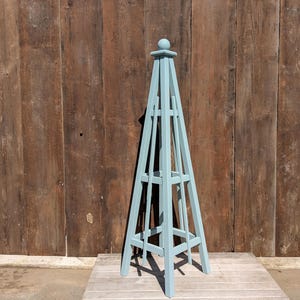 May include: A light blue wooden garden trellis in the shape of a pyramid. The trellis is standing on a wooden platform in front of a brown wooden wall.