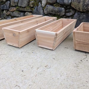 May include: Four unfinished wooden planters of varying sizes are arranged on a concrete surface. The planters are rectangular, with a natural wood grain finish. One planter has a square shape. The background features a stone wall.