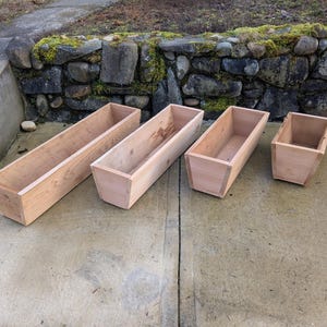 May include: Four unfinished wooden rectangular planters of varying sizes are arranged on a concrete surface. The planters are made of light-coloured wood and have a simple, open design, suitable for outdoor use.