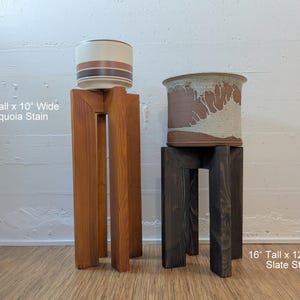 May include: Two wooden plant stands. The taller stand is 61 cm tall and 25 cm wide with a sequoia stain. The shorter stand is 41 cm tall and 30 cm wide with a slate stain. Each stand supports a ceramic pot.