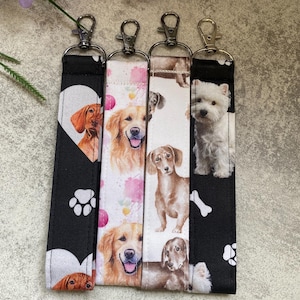 May include: Four dog-themed keychains with silver clasps. The keychains display different dog breeds, such as a Vizsla, Golden Retriever, Dachshund, and West Highland White Terrier. The keychains have black and white backgrounds with paw prints and heart designs.