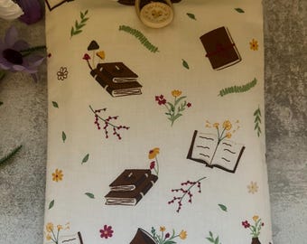 May include: Cream-colored book sleeve with a repeating pattern of brown books, flowers, and leaves. The sleeve is secured with a wooden button and loop closure. The design includes open and closed books, mushrooms, and floral accents.