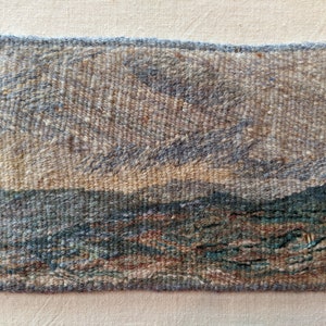 May include: A woven wall hanging depicting a landscape scene with a blue sky, brown hills, and a blue body of water. The piece is made with a variety of textures and colors.