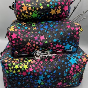 Peut inclure: Trois pochettes noires à fermeture éclair avec un motif d'étoiles arc-en-ciel. Les pochettes sont empilées les unes sur les autres. La plus grande pochette a une étiquette en acrylique transparente qui indique "Made By SharpCupcake".