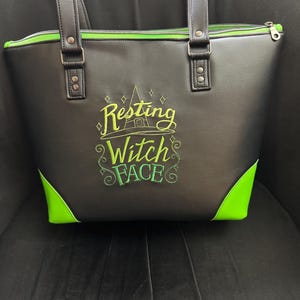 May include: Black tote bag with green accents and handles. The bag features the embroidered phrase "Resting Witch Face" in green and yellow. The bag has a zipper closure and is sitting on a dark surface.