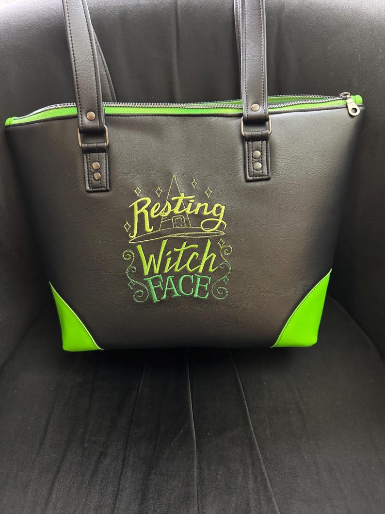 May include: Black tote bag with green accents and handles. The bag features the embroidered phrase "Resting Witch Face" in green and yellow. The bag has a zipper closure and is sitting on a dark surface.