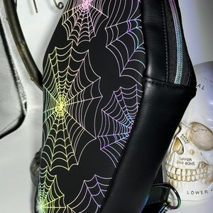 May include: Black faux leather crossbody bag with a zipper closure and an adjustable strap. The bag features a holographic spiderweb design.