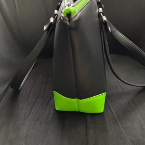 May include: Black tote bag with neon green accents on the bottom and zipper. The bag has long black straps and silver-colored hardware. The interior lining is gray. The bag is made of a smooth material.