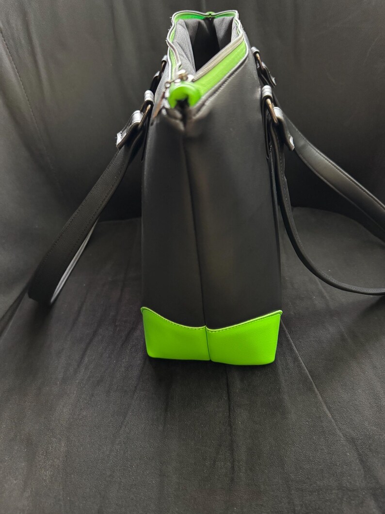 May include: Black tote bag with neon green accents on the bottom and zipper. The bag has long black straps and silver-colored hardware. The interior lining is gray. The bag is made of a smooth material.