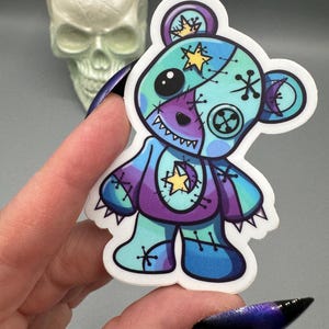 May include: A blue and purple patchwork teddy bear sticker with a stitched-on star and moon. The bear has a toothy grin and is surrounded by a white border.