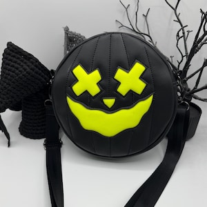 May include: Black round crossbody bag with a jack-o'-lantern face design. The bag features a stitched pumpkin shape with a neon yellow face. It has a black adjustable strap and a black bow on the side. A Halloween-themed accessory.
