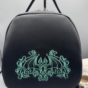 May include: Black faux leather bag with a green embroidered bat design. The bat is surrounded by a decorative floral pattern.