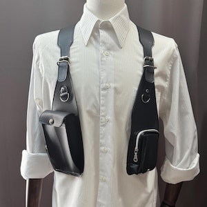 Könnte beinhalten: Schwarzes Brustgeschirr aus Leder mit zwei Taschen und verstellbaren Riemen. Das Geschirr wird über einem weißen Hemd mit Knöpfen getragen. Die Taschen haben silberfarbene Beschläge und Reißverschlüsse. Das Geschirr ist für das Tragen über der Brust konzipiert.