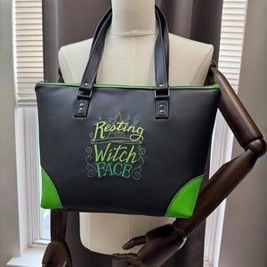 May include: Black tote bag with green accents and handles. The bag features the embroidered phrase "Resting Witch Face" in green. The corners and zipper trim are also green. The bag is displayed on a mannequin.
