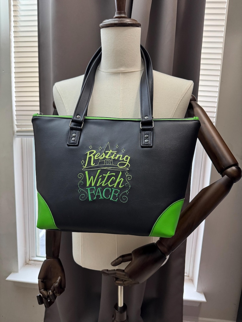 May include: Black tote bag with green accents and handles. The bag features the embroidered phrase "Resting Witch Face" in green. The corners and zipper trim are also green. The bag is displayed on a mannequin.