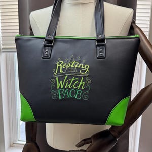 May include: Black tote bag with green accents and handles. The bag features the embroidered phrase "Resting Witch Face" in green and yellow. The bag has a zipper closure and is displayed on a mannequin.