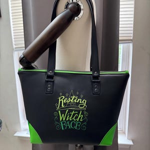 May include: Black tote bag with green accents and the phrase "Resting Witch Face" embroidered in green and yellow. The bag has black handles and a green zipper.