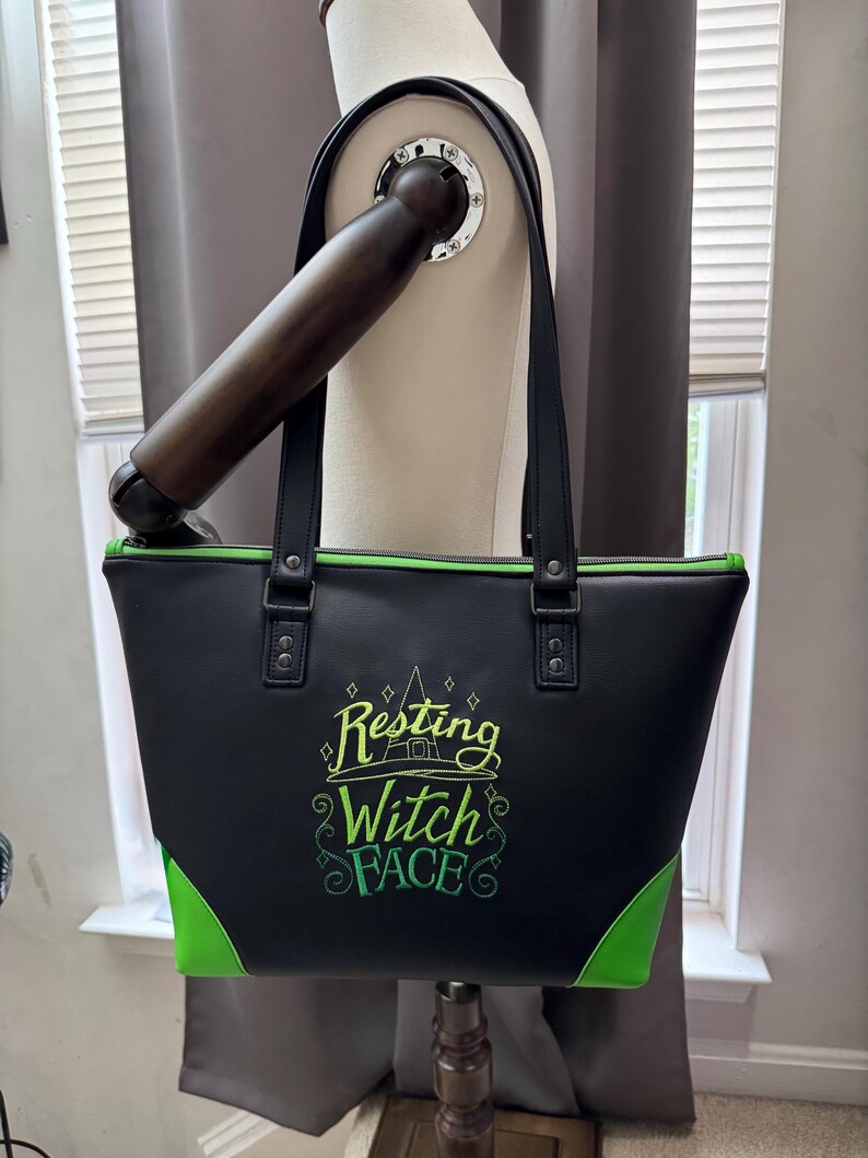May include: Black tote bag with green accents and the phrase "Resting Witch Face" embroidered in green and yellow. The bag has black handles and a green zipper.