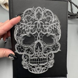 May include: Black faux leather journal with a silver embroidered skull design. The skull is intricately detailed with floral and lace-like patterns. The journal is rectangular and has a stitched border.