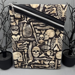 May include: A black and beige zippered pouch with a repeating pattern of human skeletal bones. The pouch has a black zip and trim, with a silver zip pull. The background is a neutral grey.