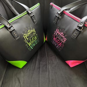 May include: Two black tote bags with the text "Resting Witch Face" in colorful lettering. One bag has green accents, the other pink. Both have black handles and zippers.