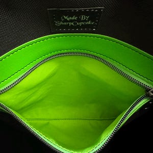 May include: Close-up of a black bag interior with a bright green lining and trim. A silver zipper runs along the opening. The text "Made By SharpCupcake" is visible on a small black tag.