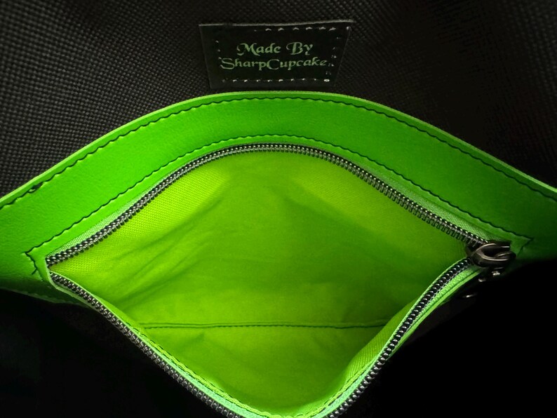 May include: Close-up of a black bag interior with a bright green lining and trim. A silver zipper runs along the opening. The text "Made By SharpCupcake" is visible on a small black tag.