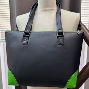 May include: Black tote bag with green accents on the corners and interior trim. The bag has two black handles and silver-colored hardware. The bag is rectangular and has a zipper closure.
