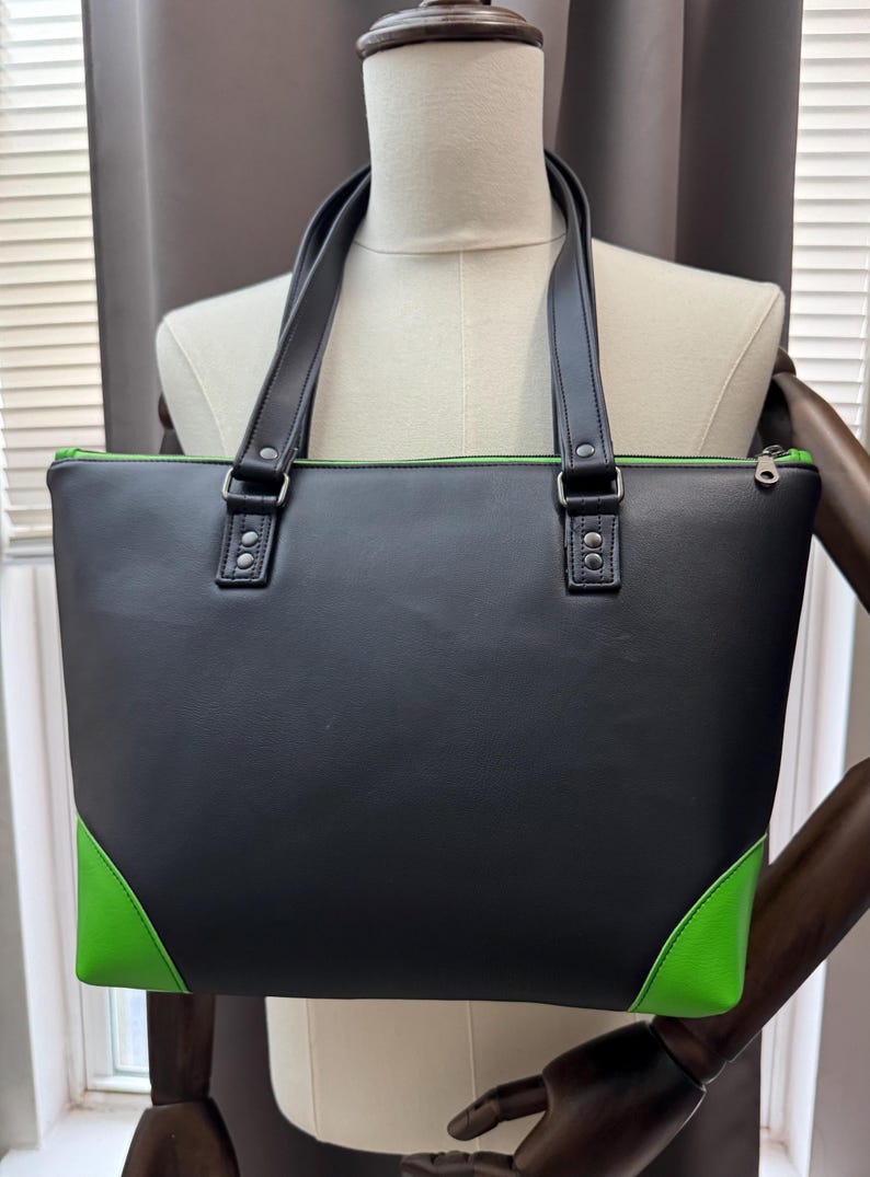 May include: Black tote bag with green accents on the corners and interior trim. The bag has two black handles and silver-colored hardware. The bag is rectangular and has a zipper closure.