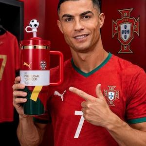 May include: A red, green, and white tumbler with a handle, featuring the text CR7 PORTUGAL TUMBLER, a Puma logo, and the Portugal national team crest. The lid includes a straw topped with a small black and white soccer ball decoration.