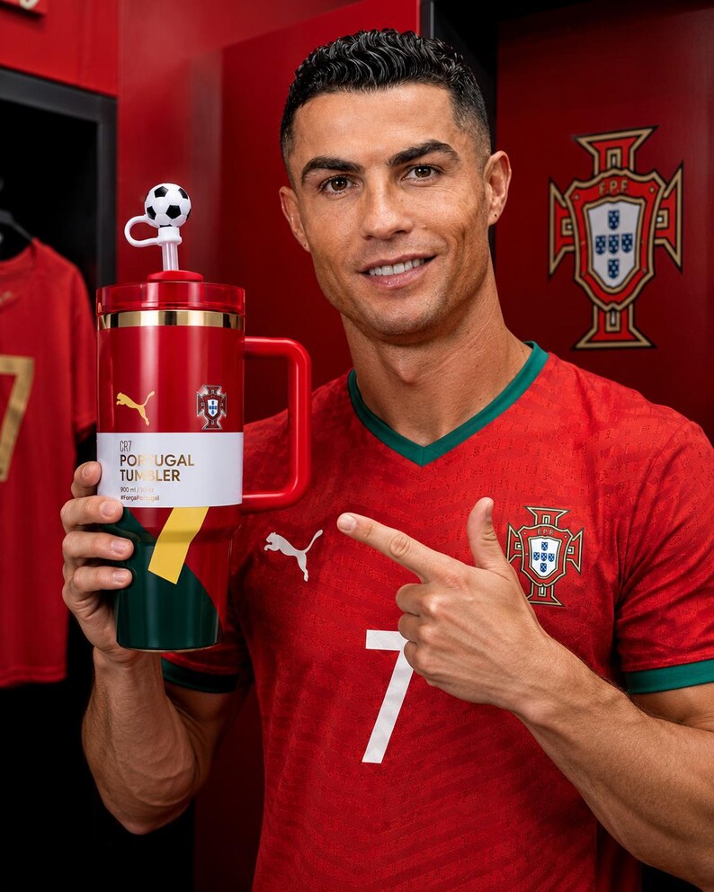 May include: A red, green, and white tumbler with a handle, featuring the text CR7 PORTUGAL TUMBLER, a Puma logo, and the Portugal national team crest. The lid includes a straw topped with a small black and white soccer ball decoration.