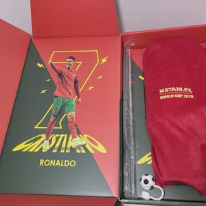 May include: A red gift box containing a Cristiano Ronaldo graphic print with a large yellow number seven, a red velvet pouch with the gold text Stanley World Cup 2026, a clear plastic straw, and a small soccer ball charm with a white loop attachment.