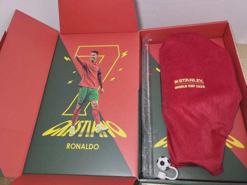 May include: A red gift box containing a Cristiano Ronaldo graphic print with a large yellow number seven, a red velvet pouch with the gold text Stanley World Cup 2026, a clear plastic straw, and a small soccer ball charm with a white loop attachment.