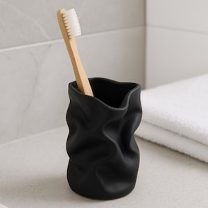 Puede incluir: Un portacepillos de cerámica negra con un diseño arrugado único. Un cepillo de dientes de madera con cerdas blancas está dentro. El soporte está sobre una encimera blanca junto a una toalla blanca doblada. Decoración de baño.