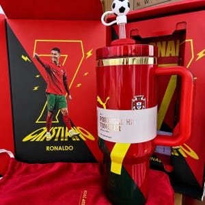 May include: A red and green tumbler with a handle, featuring a soccer ball topper on the straw and a white band labeled CR7 PORTUGAL TUMBLER 900 ml / 30 oz. The tumbler is displayed in a red box with a graphic of Cristiano Ronaldo and the number 7.