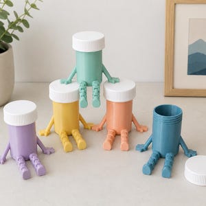 May include: A collection of five small plastic containers shaped like figures with articulated arms and legs. Four containers have white screw-on lids, while one blue container sits open without its lid. The colors include purple, yellow, green, peach, and blue.