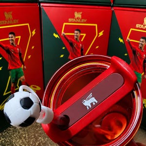 May include: A red Stanley tumbler lid with a rotating closure and a white and black soccer ball straw topper. The lid features the Stanley bear logo in white. In the background, red and green boxes display the name RONALDO and a soccer player.