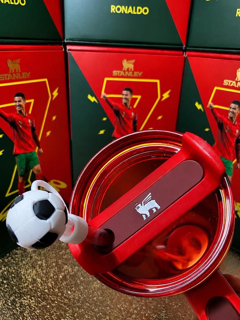 May include: A red Stanley tumbler lid with a rotating closure and a white and black soccer ball straw topper. The lid features the Stanley bear logo in white. In the background, red and green boxes display the name RONALDO and a soccer player.