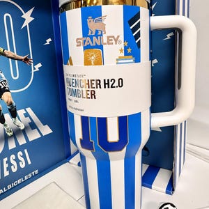 May include: A white and blue striped Stanley Quencher H2.0 tumbler featuring a gold rim, a large white handle, and a paper band with the text The Flowstate Quencher H2.0 Tumbler 40 oz 1.18 L. The tumbler displays the number 10 and a soccer ball charm.